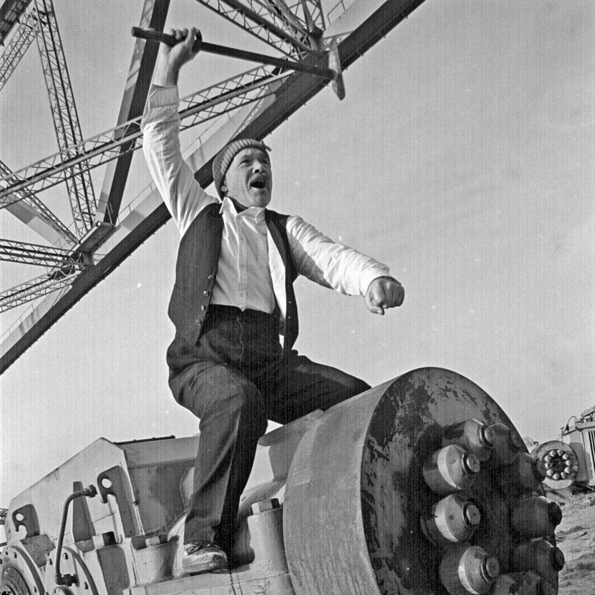 Black and white photograph of a man riding on an old piece of machinery brandishing a rusty, hammer-like tool. The man is wearing a white shirt with a black vest, black trousers and a grey wooly hat.