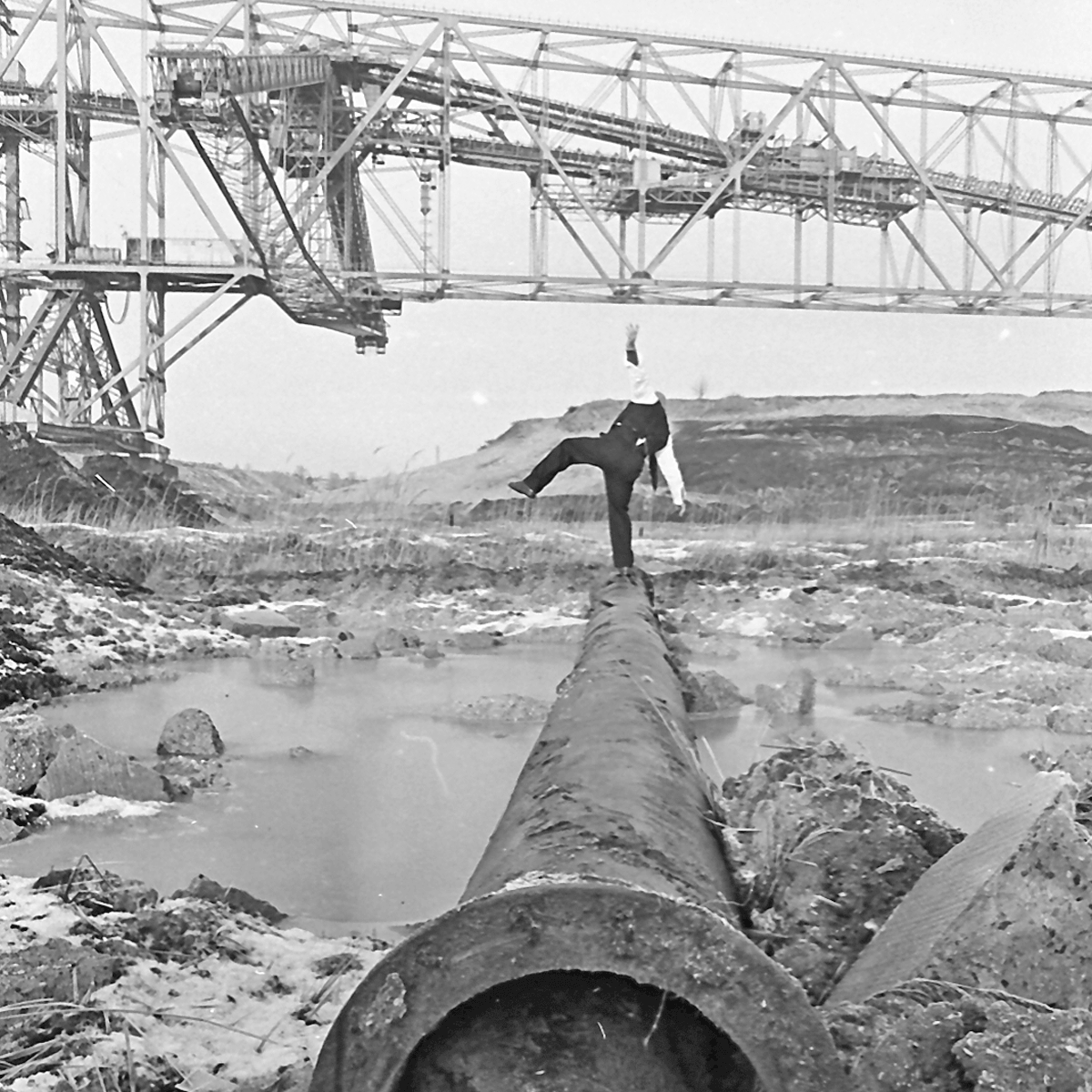 Black and white photograph of a man balancing on an old rusty pipe. The man is wearing a white shirt with a black vest, black trousers and a grey wooly hat.